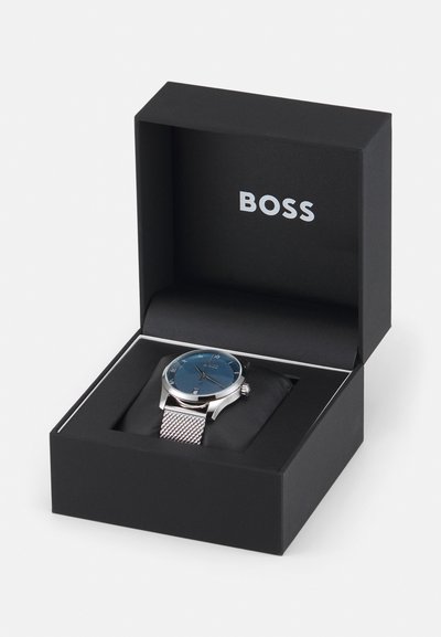Montre en acier inoxydable argenté avec un cadran texturé bleu, entourée d'un bracelet en maille, présentée dans une boîte noire avec le logo "BOSS".