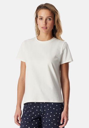 Jonge vrouw met golvend blond haar, gekleed in een effen wit T-shirt met korte mouwen en marineblauwe broek met een klein wit stippenpatroon.