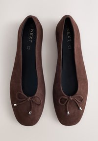 Paire de ballerines en daim marron foncé avec des bouts arrondis et de petits nœuds à l'avant, vues de dessus sur un fond neutre.