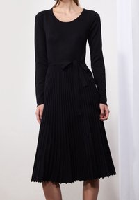 Robe noire plissée avec manches longues, encolure ronde et ceinture nouée à la taille. Confectionnée dans un tissu à texture lisse. Détail ourlet festonné.