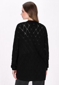 Cardigan negro de punto con un patrón geométrico de calados y puños de canalé. Mangas largas y un corte relajado. La vista trasera muestra el escote.