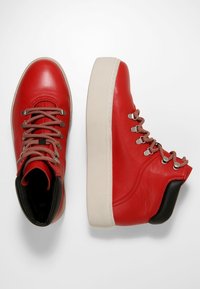 Czerwone skórzane buty typu high-top z beżową podeszwą, z brązowymi i czerwonymi sznurówkami, metalowymi oczkami oraz czarnym akcentem na pięcie.