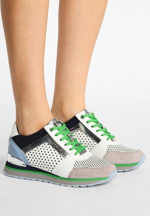 Fødder med hvide sneakers med sorte stjernemønstre, grønne snørebånd, lyseblå accenter og såler med grønne striber på ensfarvet baggrund.