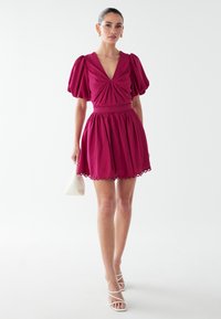 Fuchsia Kleid mit Puffärmeln, tiefem V-Ausschnitt, gerafftem Oberteil und Spitzenbesatz am Saum. Kombiniert mit weißen, schmalen Absätzen und einer kleinen Tasche.