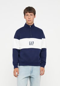 Svetrová mikina v námorníckej a bielej farbe s farebnými blokmi, vysokým golierom, štvrťovým zipsom a logom "GAP" na prednej strane. Mäkka textúra a voľný strih.