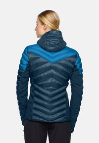 Donna vista da dietro che indossa un giubbotto imbottito trapuntato di colore blu e verde scuro con cappuccio, in piedi su uno sfondo chiaro e uniforme.