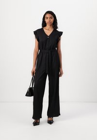 Combinaison noire avec des manches courtes, un front à boutons, une taille ceinturée et des jambes larges. Le mannequin tient un petit sac noir et porte des chaussures pointues noires.