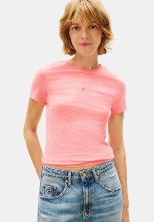 Giovane donna che indossa una t-shirt rosa "Tommy Jeans" e jeans in denim blu a vita alta, in piedi con le mani nelle tasche.