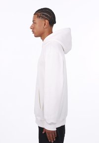 Hvid hætte sweatshirt med kengurulomme, tekstureret stof og ribbede manchetter. Sidevisning der viser minimalistisk design og subtile detaljer.