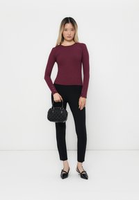 Una donna che indossa una maglietta attillata a maniche lunghe bordeaux, pantaloni neri slim, tacchi a punta neri, mentre tiene una piccola borsa nera con texture.