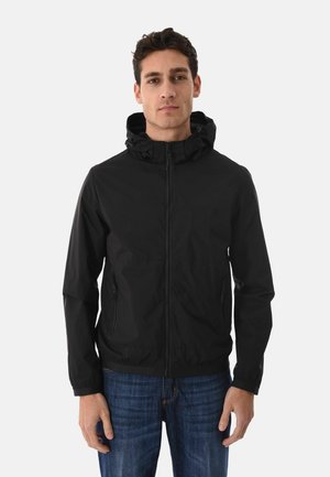 Giovane uomo che indossa una giacca nera con cappuccio e cerniera, con tasche laterali, e jeans blu, in piedi davanti a uno sfondo chiaro e uniforme.