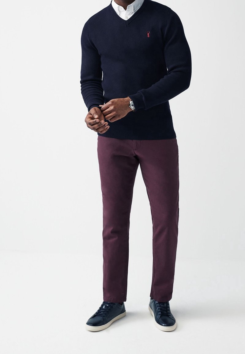 Merimies v-kaula-aukollinen villapaita, viininpunaiset housut ja tummat lenkkarit. Rakenteinen kangas, kapea istuvuus, ja hienovarainen logotunnus rinnassa.