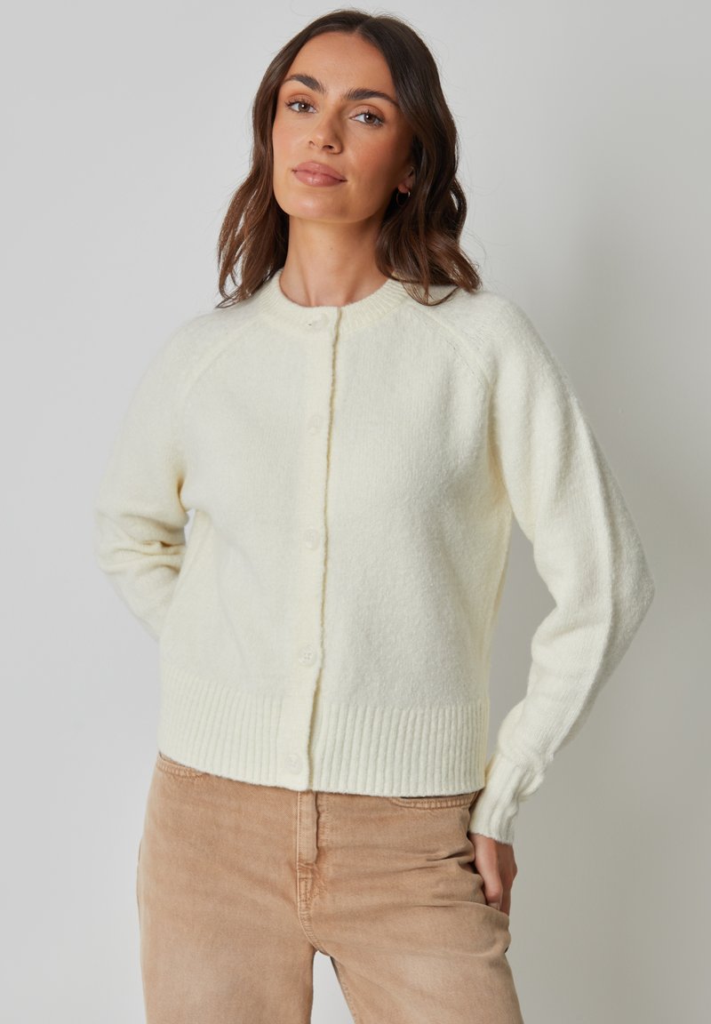 Weißer, flauschiger Cardigan mit rundem Ausschnitt, Knopfleiste, geripptem Saum und Bündchen. Einfaches Design mit Raglanärmeln. Weiche Textur.