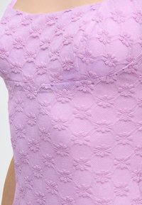 Κοντινό πλάνο σε ύφασμα από λιλά δαντέλα με μικρά ανθικά μοτίβα σε εφαρμοστό ένδυμα, που αναδεικνύει λεπτομέρειες υφής και διαφανείς περιοχές.