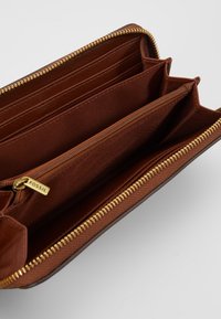 Portefeuille en cuir marron avec fermeture éclair, comprenant plusieurs emplacements pour cartes, un compartiment zippé et un logo en relief. Intérieur texturé.
