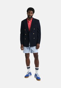 Blazer de doble botonadura en navy a rayas con botones dorados, polo rojo, pantalones cortos azul claro, zapatillas azules con rayas blancas y calcetines a rayas.