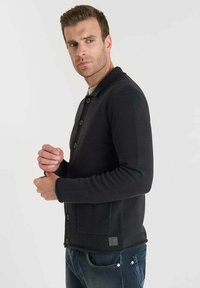 Uomo che regola la manica di un cardigan nero abbottonato con tasca a toppa, indossando jeans scuri, davanti a uno sfondo chiaro e uniforme.
