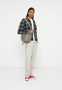 Borsa a tracolla in tessuto grigio con cinturino, camicia a quadri nera e verde sopra una T-shirt bianca, pantaloni beige chiaro e sneakers rosse.