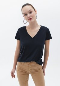 Černé krátké tričko s výstřihem do V vyrobené z jemného materiálu, v kombinaci s světle béžovými úzkými kalhotami. Model stojí na jednobarevném bílém pozadí.