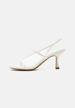 Sandale albe de damă cu toc, cu vârf decupat, barete subțiri în cruce și o curea tip slingback pe un fundal deschis.