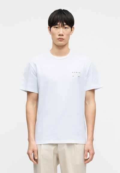 T-shirt en coton blanc à manches courtes, col rond, détail logo noir minimaliste sur la poitrine gauche, ourlet droit. Porté avec un pantalon beige clair.