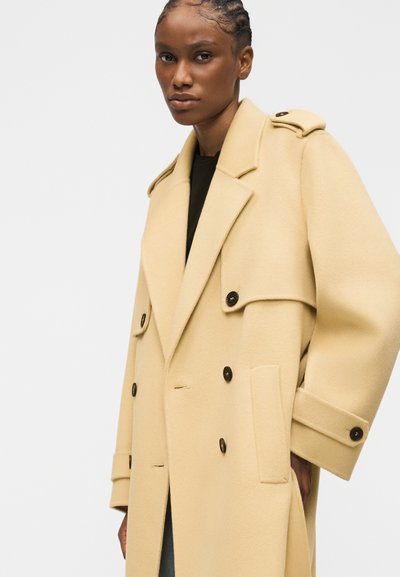 Γυναίκα με πλεγμένα μαλλιά που φοράει υπερμέγεθες μπεζ παλτό με διπλό στήθος, μαύρα κουμπιά και πλαϊνές τσέπες με καπάκι, στέκεται μπροστά σε απλό φόντο.