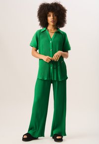 Green pleated co-ord set featuring a short-sleeve button-up shirt and wide-leg pants, crafted from a soft, textured fabric with a relaxed fit.