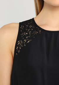 Czarny top bez rękawów z koronkowym zdobieniem na ramieniu i dekolcie, wykonany z gładkiego materiału na całej długości.
