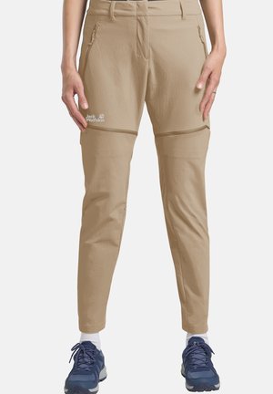 Pantaloni outdoor beige con zip staccabile, indossati con scarpe da trekking blu e calzini bianchi, vista frontale che mostra dettagli della zip e il logo Jack Wolfskin.