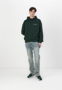 Hanorac supradimensionat de culoare verde închis, cu buzunar frontal și text alb "nu risipi cultura", asortat cu blugi de culoare bleu deschis și adidași verzi închiși.