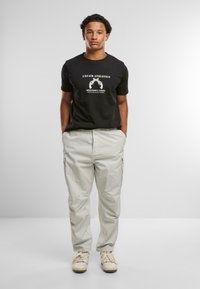 T-shirt nera in cotone con una stampa grafica bianca, abbinata a pantaloni cargo grigi chiari con tasche laterali e polsini elasticizzati, e sneakers beige.