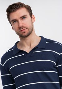 Muž s krátkými hnědými vlasy a vousy, oblečený v námořnicky modrém polo tričku s bílými vodorovnými pruhy, na čistém světlém pozadí.