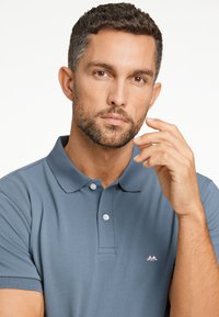 Grå polo med texturerad tyg, korta ärmar och en treknappslåsning. Har en liten vit bergslogotyp på bröstet.