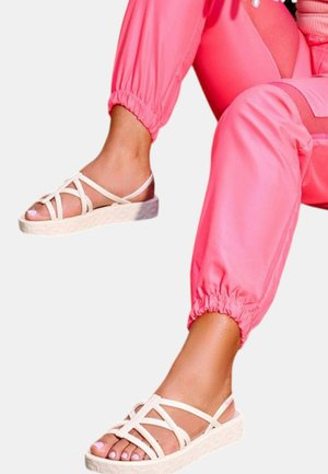 Sandali beige con cinturini incrociati e suola texturizzata, indossati con joggers rosa a manica elastica.