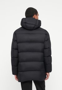 Giacca a vento nera con cappuccio, caratterizzata da un design trapuntato e una texture liscia e opaca. Il retro è leggermente più lungo con un taglio dritto.