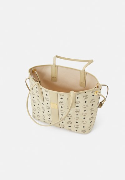 Sac à tote beige avec un extérieur à motifs présentant des logos noirs. Forme arrondie, en cuir, avec deux poignées et un intérieur ouvert.
