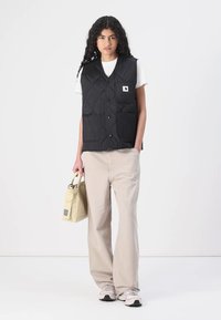 Gilet nero trapuntato con motivi rotondi, tasche frontali e patch logo, indossato sopra una maglietta bianca, abbinato a pantaloni beige e una borsa leggera.