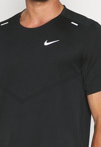 Camiseta atlética negra de manga corta, hecha de tela suave; presenta un pequeño logo blanco de Nike y dos acentos blancos en los hombros.