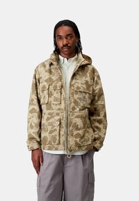 Giacca beige con motivo mimetico, dotata di cappuccio, numerose tasche frontali e chiusure a scatto. Indossata sopra una camicia leggera, con zip e orlo regolabile.