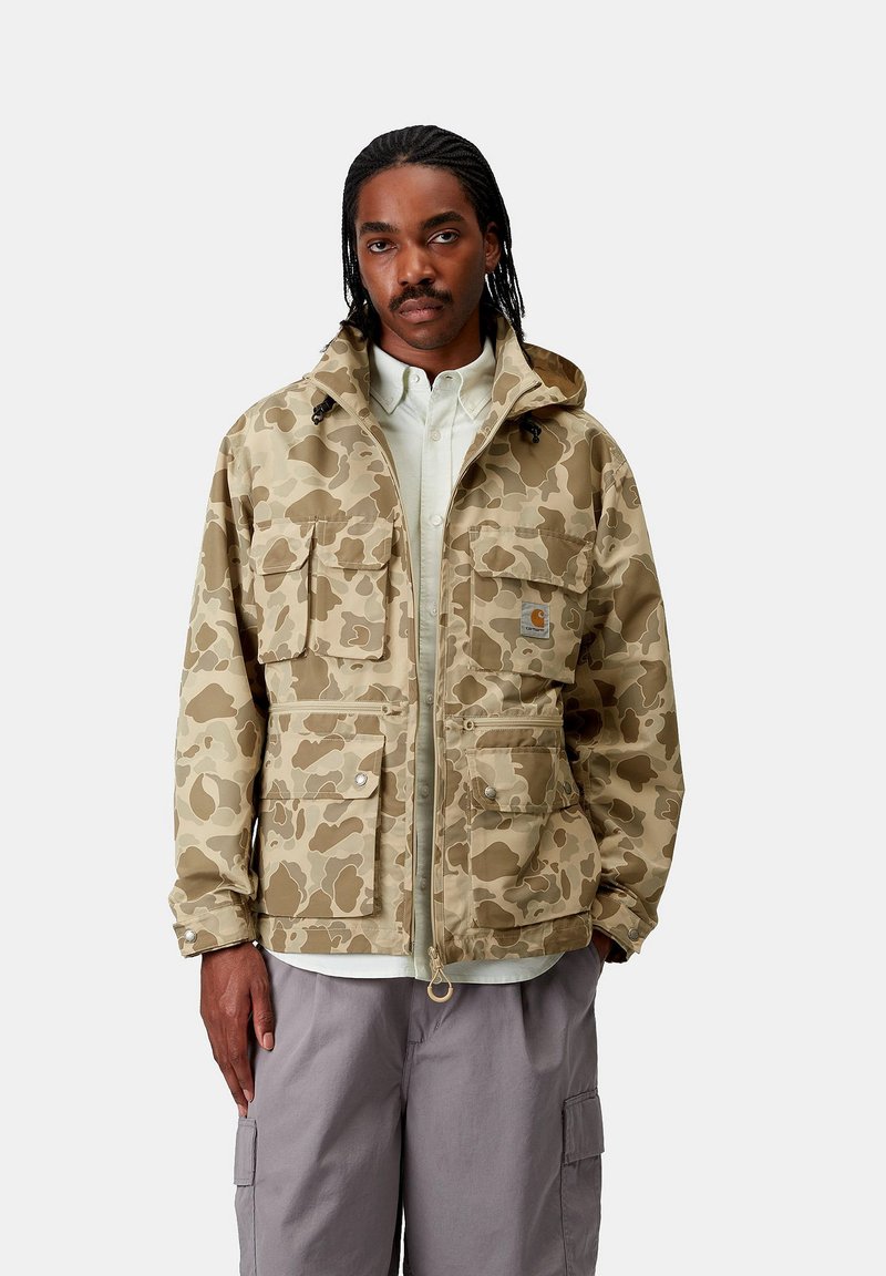 Giacca beige con motivo mimetico, dotata di cappuccio, numerose tasche frontali e chiusure a scatto. Indossata sopra una camicia leggera, con zip e orlo regolabile.
