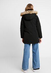 Parka noire avec capuche en fausse fourrure, dotée d'un cordon de serrage à la taille et de manches longues, associée à un jean large bleu clair et des baskets.
