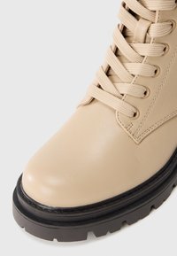 Botine din piele bej, cu vârf rotund, șireturi în față și talpă din cauciuc negru cu un model texturat de aderență. Detalii cusute pe partea superioară.