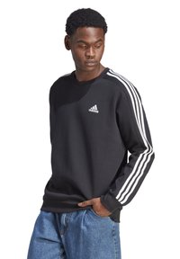 Sweatshirt preto com gola redonda, apresentando três riscas brancas em cada manga e um pequeno logótipo branco no peito. O tecido parece suave.