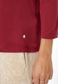 Primer plano de una mano que lleva un anillo de plata junto a una camisa de manga larga roja y una falda beige con estampado.