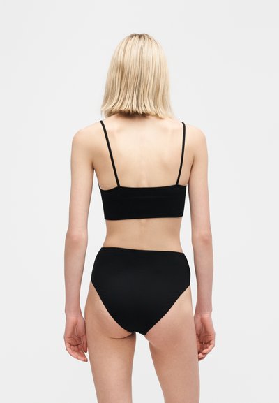 Ξανθιά γυναίκα που στέκεται γυρισμένη πλάτη, φοράει μαύρο κομμένο τοπ με λεπτές τιράντες και μαύρο ψηλόμεσο σορτς, μπροστά σε απλό φόντο.