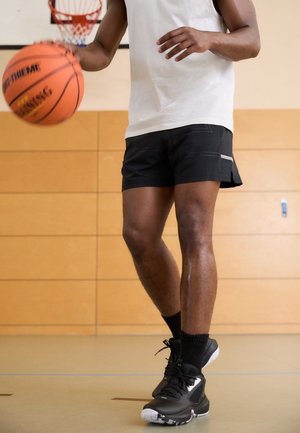 Personne en short noir et baskets dribblant un ballon de basket sur un terrain intérieur avec un panier et un mur en panneaux de bois visibles.