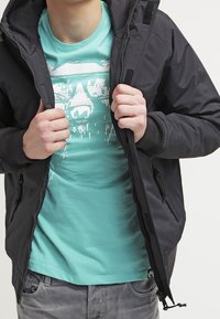 Giacca nera con zip e cappuccio, dotata di due tasche laterali. Sotto, una t-shirt grafica turchese con una stampa di montagna bianca. Pantaloni grigi.