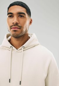 Sudadera color beige claro hecha de tela suave, con un bolsillo tipo canguro, capucha con cordón y acentos metálicos en contraste en los cordones.
