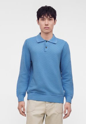 Jeune homme portant un polo texturé bleu clair à manches longues et un pantalon beige, debout devant un fond blanc uni.