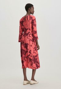 Vestido rojo con estampado floral, con mangas tres cuartos, cintura elástica, largo hasta la mitad de la pantorrilla y detalle en la espalda tipo ojal. Llevado con zapatos beige.